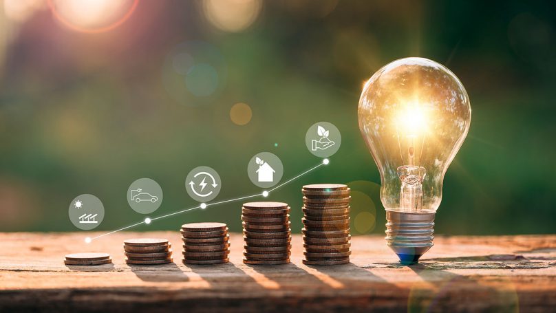  lâmpada acesa ao lado de pilhas de moedas em ordem crescente, simbolizando economia